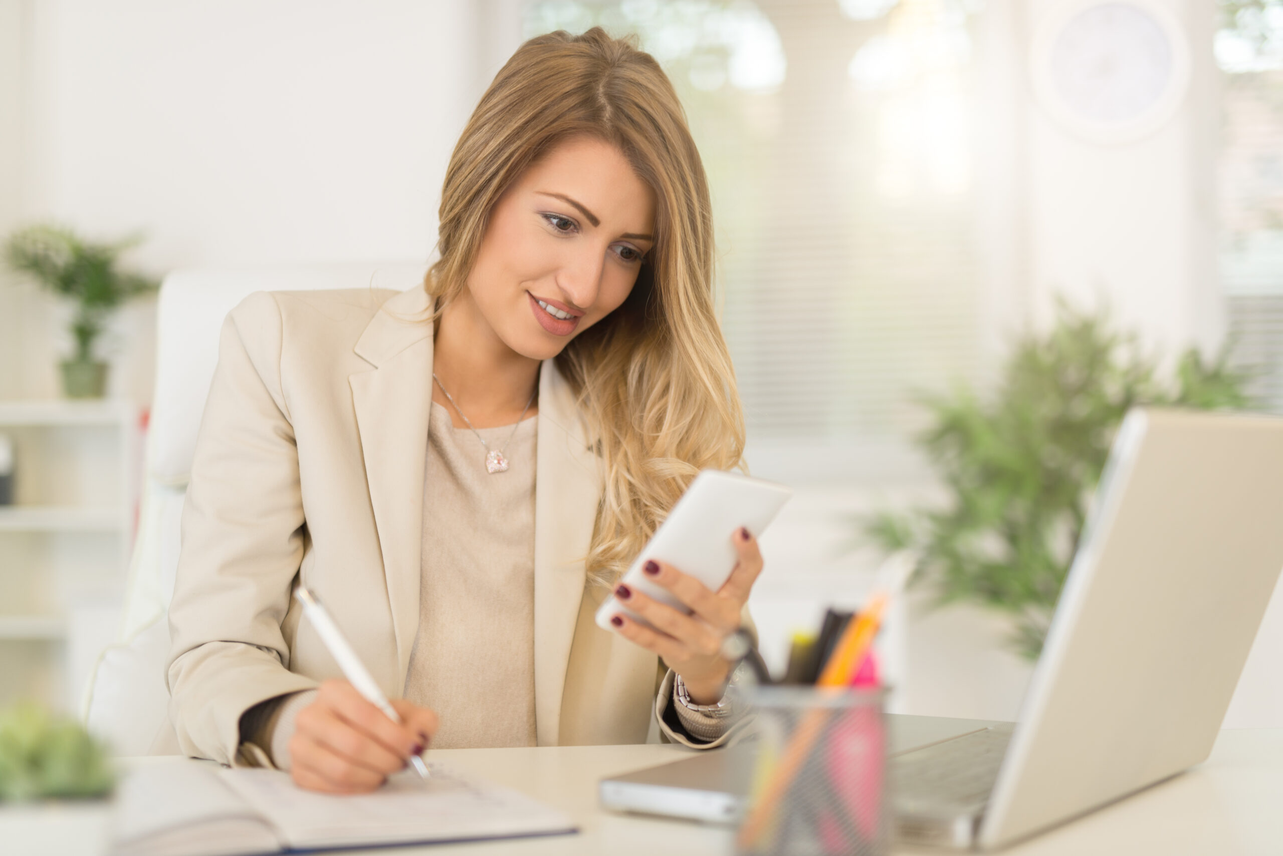 Beautiful young businesswoman sitting in the office, using phone and writing in planner.